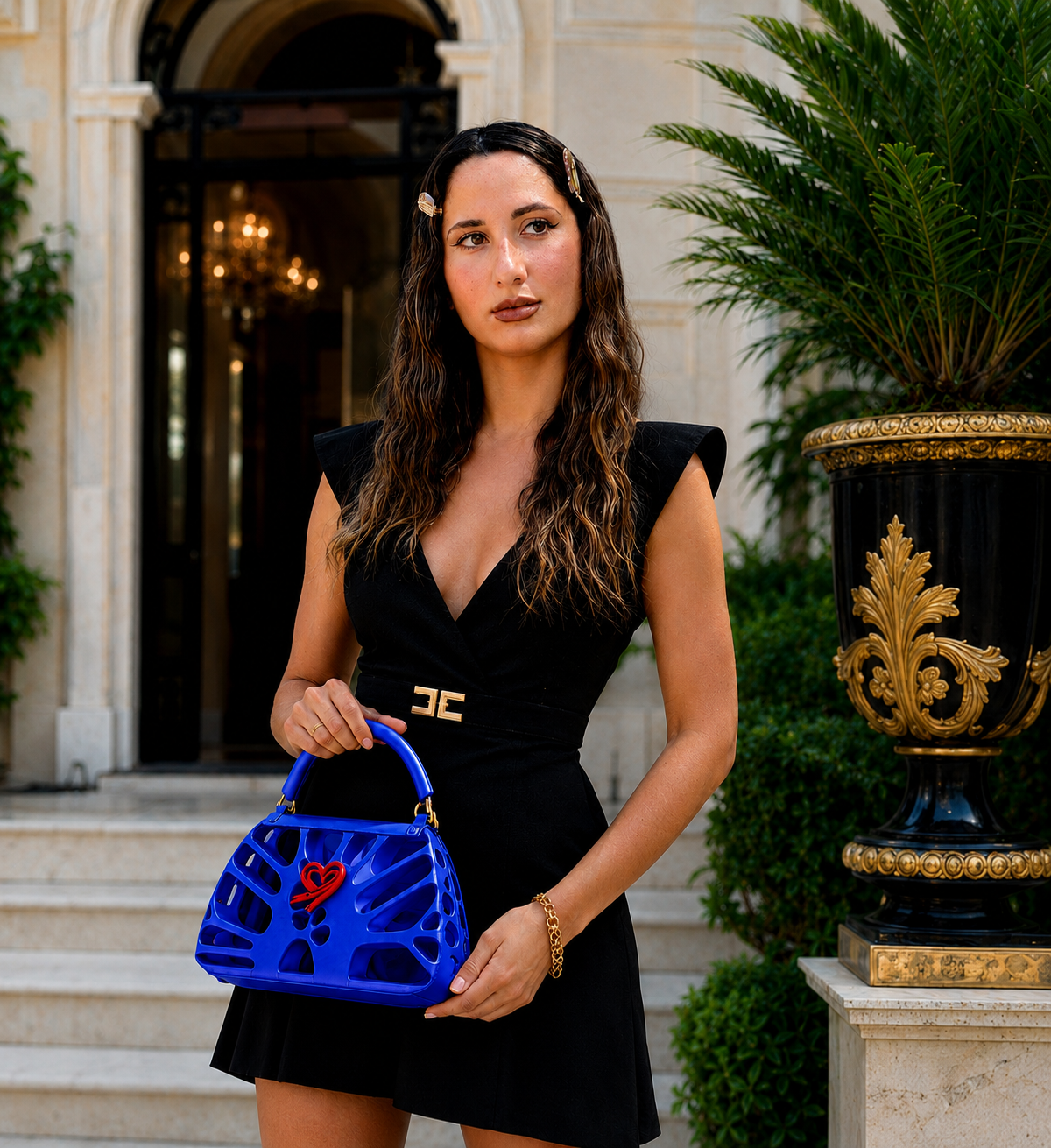 Fjoccola FDR blue women’s handbag, a 3D printed PLA Silk designer bag with sculptural openwork design, top handle and signature red heart detail, styled with an elegant black dress.
